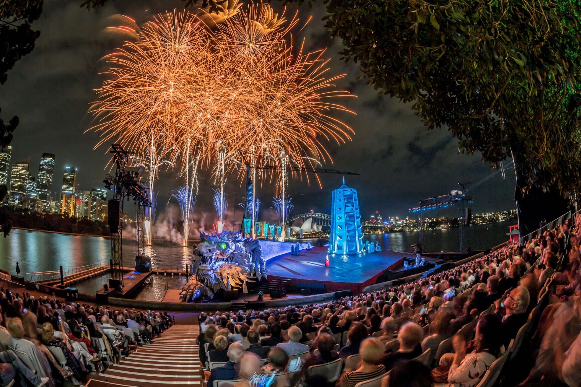 An audience of 3000 sit in an outdoor stadium on the edge of Sydney Harbour looking over a stage in front of the Sydney Opera House with performers and fireworks.