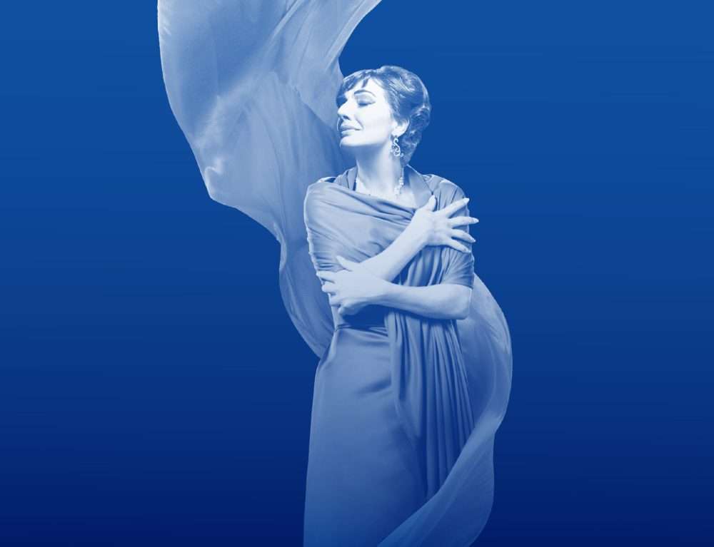 Opera Singer Maria Callas with her eyes closed and her arms wrapped around her. She's wearing a blue dress with sparkling earrings and a sparkling necklace. She's against a blue backdrop.